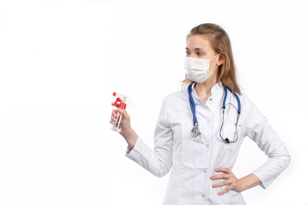 Foto gratuita una vista frontal joven doctora en traje médico blanco con estetoscopio con máscara protectora blanca posando sosteniendo spray sobre el blanco