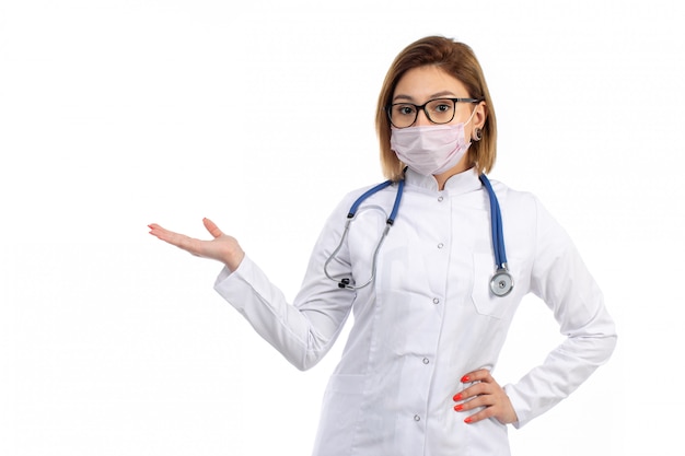 Una vista frontal joven doctora en traje médico blanco con estetoscopio con máscara protectora blanca sobre el blanco