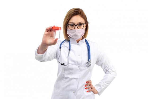 Una vista frontal joven doctora en traje médico blanco con estetoscopio con máscara protectora blanca sosteniendo el matraz sobre el blanco