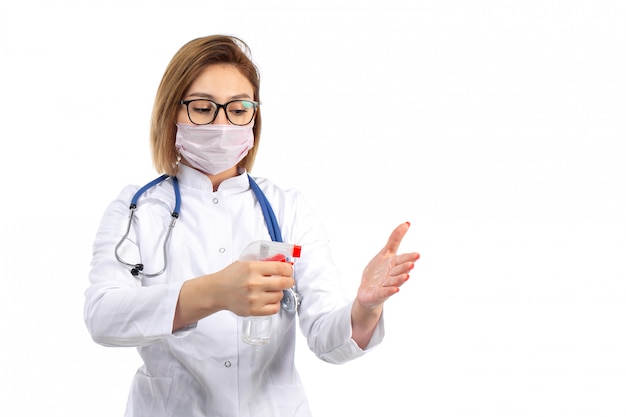 Una vista frontal joven doctora en traje médico blanco con estetoscopio con máscara protectora blanca con spray sobre el blanco