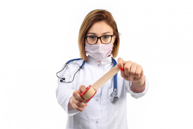 Foto gratuita una vista frontal joven doctora en traje médico blanco con estetoscopio con máscara protectora blanca con vendaje en el blanco