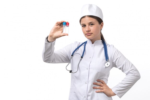 Una vista frontal joven doctora en traje médico blanco y gorra blanca con estetoscopio azul con frascos