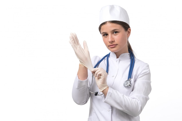 Una vista frontal joven doctora en traje médico blanco y gorra blanca con estetoscopio azul con guantes