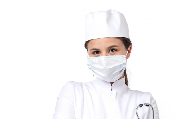 Una vista frontal joven doctora en traje médico blanco y gorra blanca con estetoscopio azul con una máscara