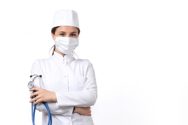 Una vista frontal joven doctora en traje médico blanco y gorra blanca con estetoscopio azul con una máscara