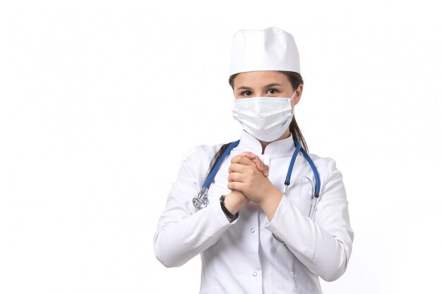 Una vista frontal joven doctora en traje médico blanco y gorra blanca con estetoscopio azul con una máscara