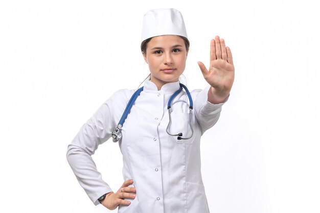 Una vista frontal joven doctora en traje médico blanco y gorra blanca con estetoscopio azul mostrando señal de stop