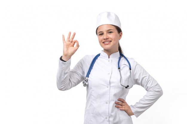 Una vista frontal joven doctora en traje médico blanco y gorra blanca con estetoscopio azul sonriendo