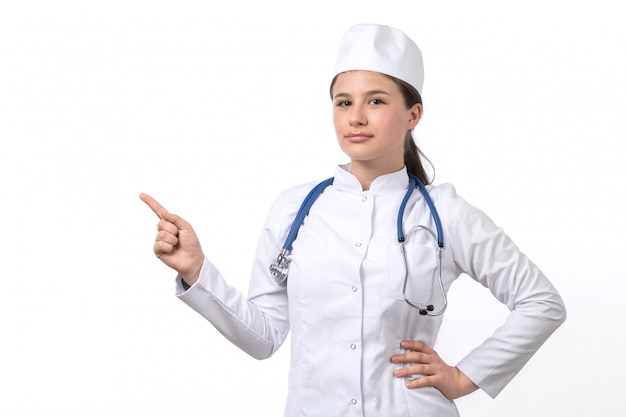 Una vista frontal joven doctora en traje médico blanco y gorra blanca con estetoscopio azul