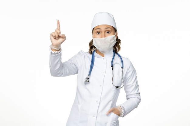 Vista frontal joven doctora en traje médico blanco y con máscara debido al coronavirus en el escritorio blanco claro