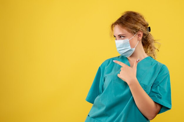 Vista frontal de la joven doctora en traje médico y máscara estéril en la pared amarilla