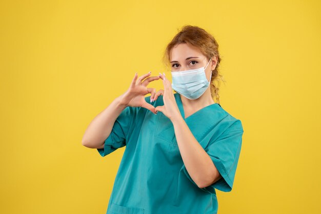 Vista frontal de la joven doctora en traje médico y máscara estéril que muestra el signo de amor en la pared amarilla