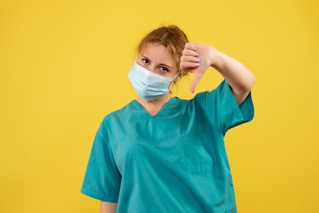 Vista frontal de la joven doctora en traje médico y máscara en la pared amarilla