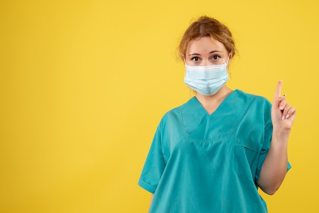 Vista frontal de la joven doctora en traje médico y máscara en la pared amarilla