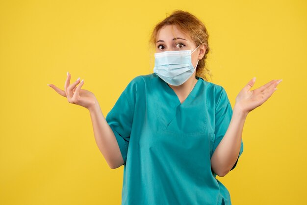 Vista frontal de la joven doctora en traje médico y máscara en la pared amarilla