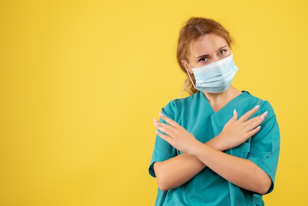 Vista frontal de la joven doctora en traje médico y máscara en la pared amarilla