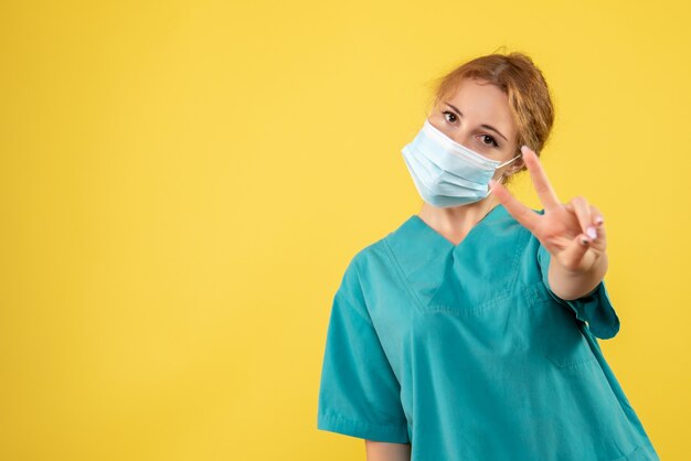 Vista frontal de la joven doctora en traje médico y máscara en la pared amarilla