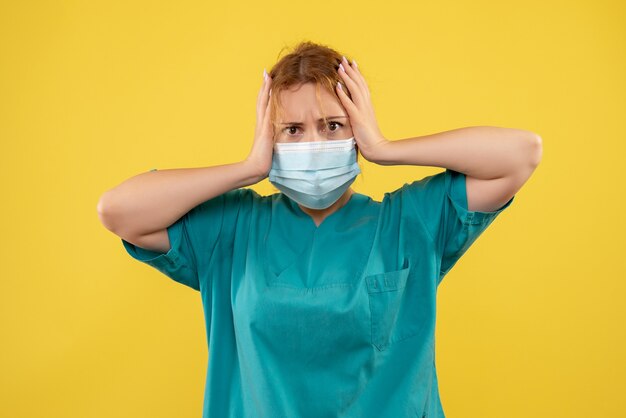 Vista frontal de la joven doctora en traje médico y máscara en la pared amarilla