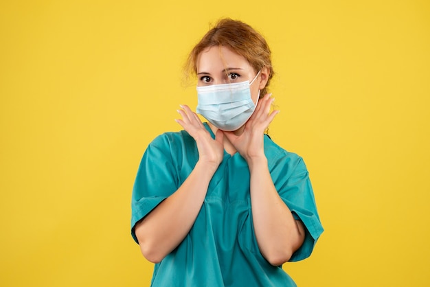 Vista frontal de la joven doctora en traje médico y máscara en la pared amarilla
