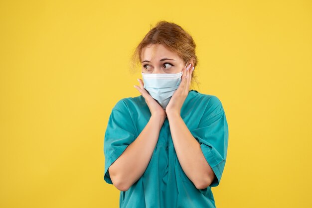 Vista frontal de la joven doctora en traje médico y máscara en la pared amarilla