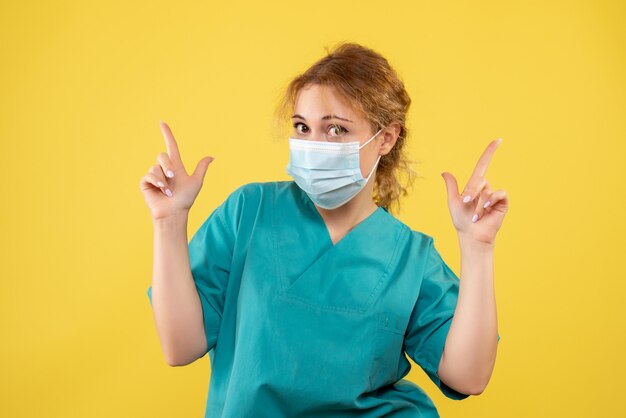 Vista frontal de la joven doctora en traje médico y máscara en la pared amarilla