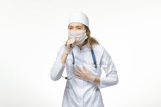 Vista frontal joven doctora en traje médico con máscara protectora debido a la tos del coronavirus en la superficie blanca