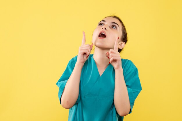 Vista frontal de la joven doctora en traje médico mirando hacia arriba en la pared amarilla