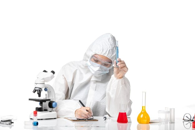 Vista frontal joven doctora en traje de protección blanco con máscara debido a covid sosteniendo una solución azul escribiendo sobre fondo blanco covid pandemia de salpicaduras- virus