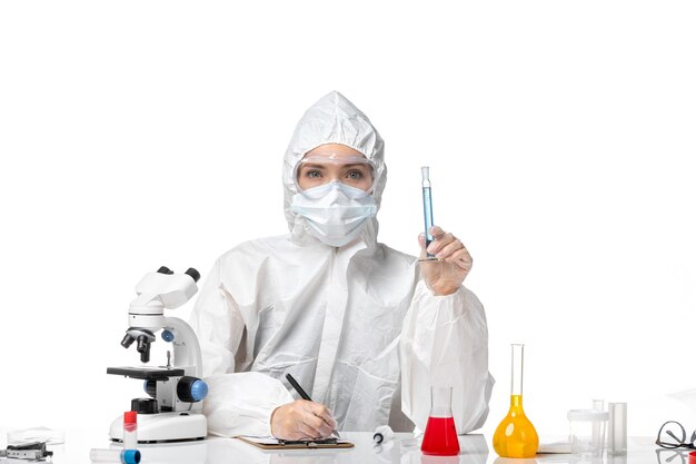 Vista frontal joven doctora en traje de protección blanco con máscara debido a covid sosteniendo una solución azul sobre fondo blanco claro splash pandemia covid- virus