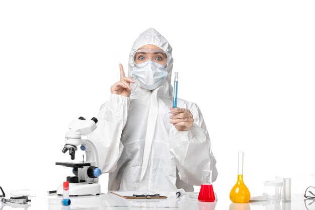 Vista frontal joven doctora en traje de protección blanco con máscara debido a covid sosteniendo una solución azul sobre fondo blanco covid pandemia de salpicaduras- virus