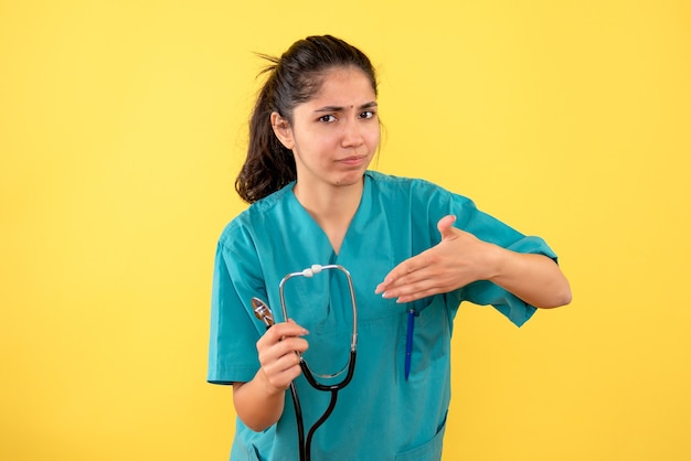 Vista frontal de la joven doctora en uniforme apuntando al estetoscopio de pie en la pared amarilla