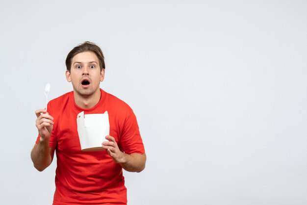 Vista frontal del joven emocional conmocionado en blusa roja con caja de papel sobre fondo blanco.
