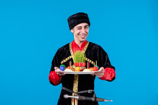 Vista frontal de un joven emotivo sonriente vestido con un vestido tradicional que sostiene una bandeja llena de confitería nacional de fondo azul