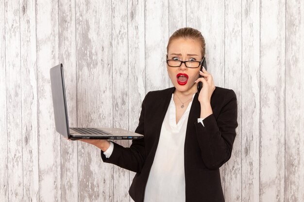 Vista frontal joven empresaria en ropa estricta chaqueta negra usando su computadora portátil y hablando por teléfono en la superficie blanca