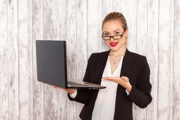 Vista frontal joven empresaria en ropa estricta chaqueta negra usando su computadora portátil con una leve sonrisa en la pared blanca trabajo trabajo oficina mujer trabajadora de negocios