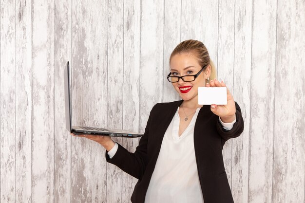 Vista frontal joven empresaria en ropa estricta chaqueta negra usando su computadora portátil y sosteniendo la tarjeta en la superficie blanca