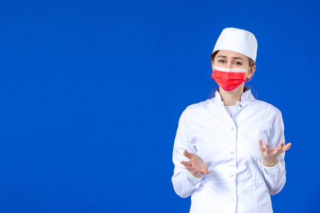 Vista frontal de la joven enfermera estresada en traje médico con máscara roja sobre el azul