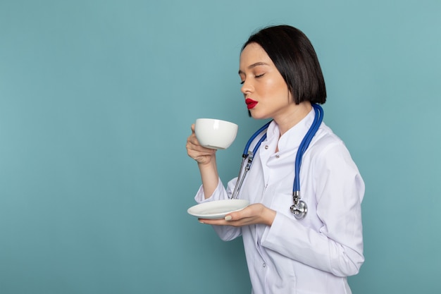 Una vista frontal joven enfermera en traje médico blanco y estetoscopio azul bebiendo té