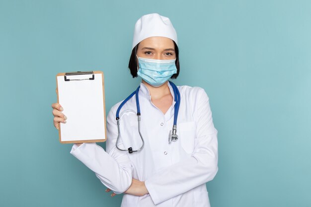 Una vista frontal joven enfermera en traje médico blanco y estetoscopio azul con bloc de notas en la máscara