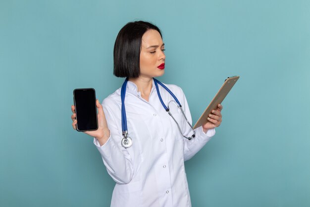 Una vista frontal joven enfermera en traje médico blanco y estetoscopio azul con bloc de notas y teléfono