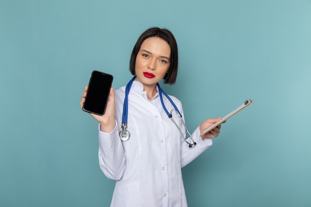 Una vista frontal joven enfermera en traje médico blanco y estetoscopio azul con bloc de notas y teléfono