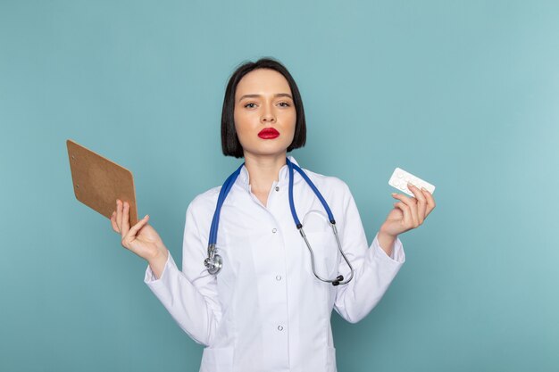 Una vista frontal joven enfermera en traje médico blanco y estetoscopio azul con bloc de notas