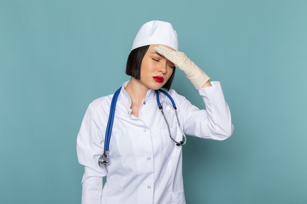 Una vista frontal joven enfermera en traje médico blanco y estetoscopio azul con dolor de cabeza