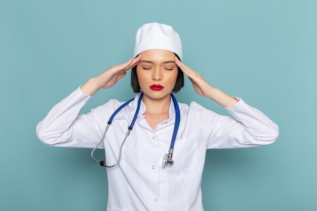 Una vista frontal joven enfermera en traje médico blanco y estetoscopio azul con dolor de cabeza