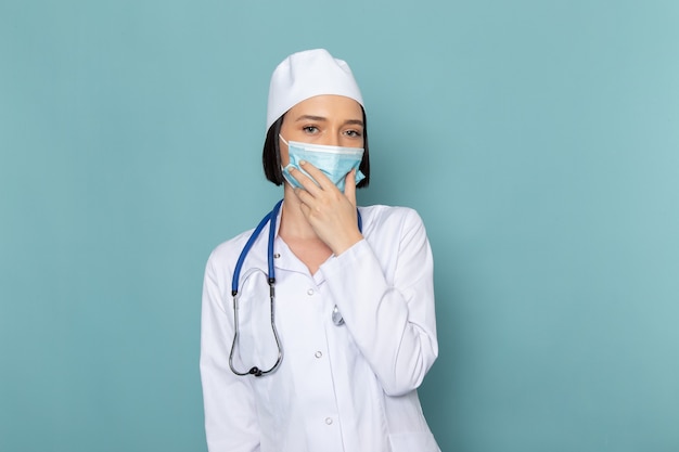 Una vista frontal joven enfermera en traje médico blanco y estetoscopio azul con máscara en el escritorio azul médico médico del hospital