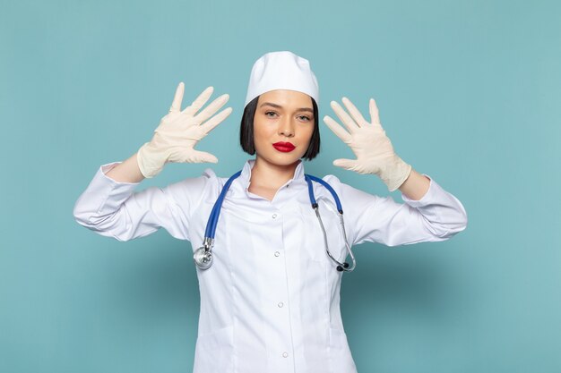 Una vista frontal joven enfermera en traje médico blanco y estetoscopio azul mostrando sus manos en el escritorio azul médico médico del hospital