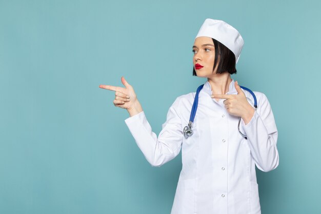 Una vista frontal joven enfermera en traje médico blanco y estetoscopio azul posando en el escritorio azul médico médico del hospital