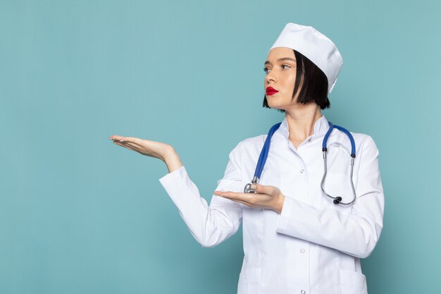 Una vista frontal joven enfermera en traje médico blanco y estetoscopio azul posando en el escritorio azul médico médico del hospital