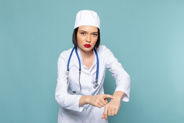 Una vista frontal joven enfermera en traje médico blanco y estetoscopio azul posando en el escritorio azul médico médico del hospital