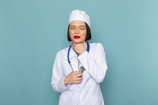 Una vista frontal joven enfermera en traje médico blanco y estetoscopio azul con problemas de garganta